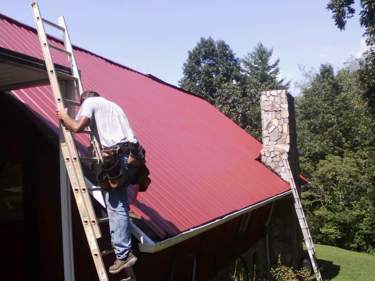 Roofer on a ladder beside a red metal roof and stone chimney — Soffit & Fascia Repair by Roofing Brothers in Tarentum